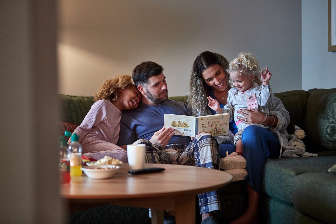 Family cuddles on a sofa, reading picture books; a toddler flashes peace signs. Coffee table holds popcorn, drinks, mug. Text visible: A PETER RABBIT, Three Little Bunnies.